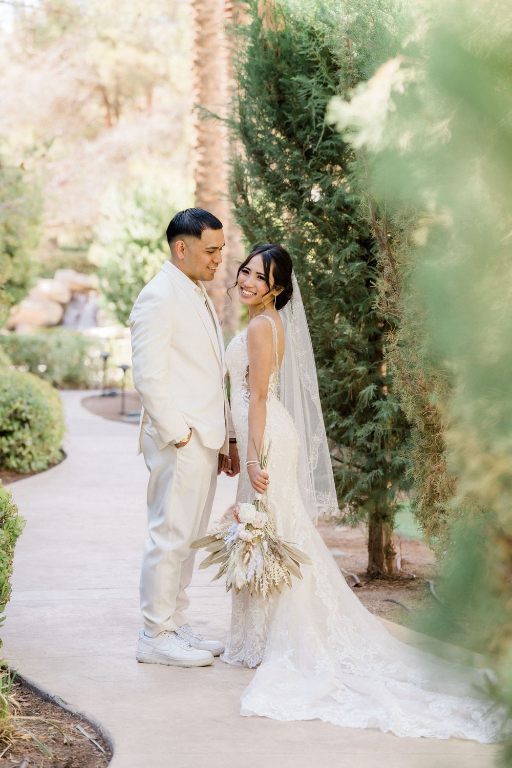 Newlyweds share joyful moment on garden path in coordinated cream outfits.