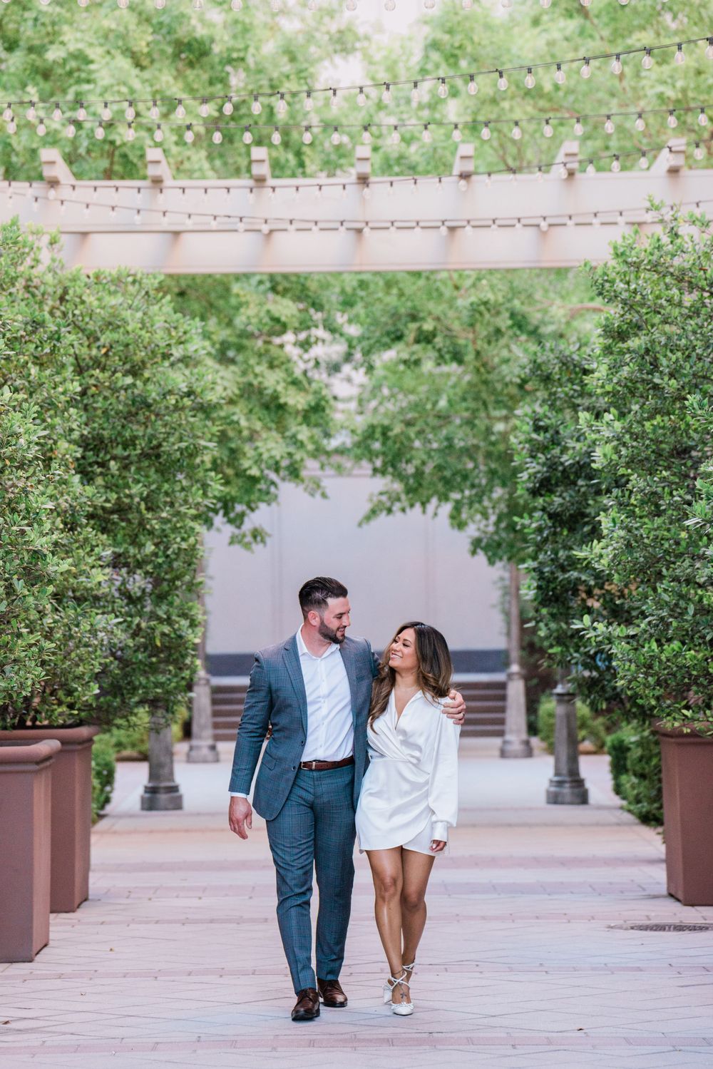A couple strolls beneath string lights and greenery in an urban courtyard setting.