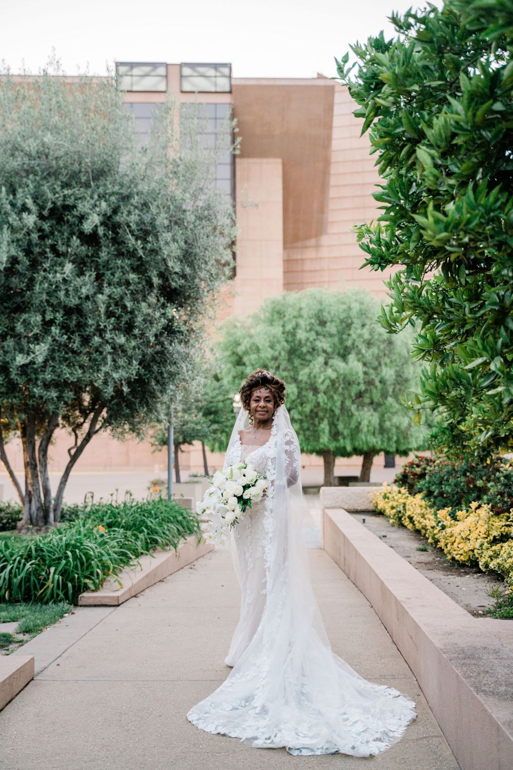 Bride in lace gown poses along garden pathway with architectural elements in background.