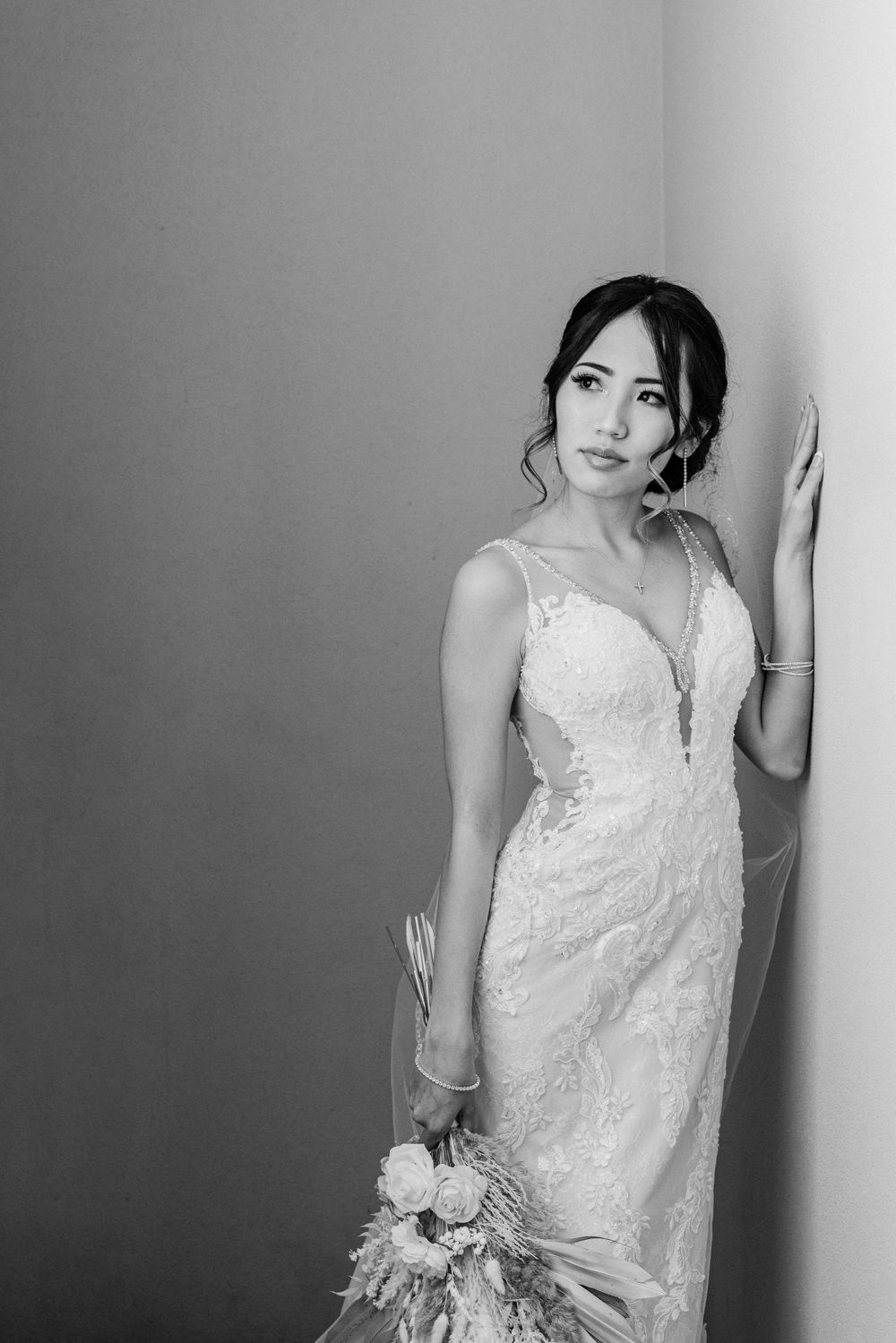 A bride in a lace wedding gown poses elegantly against a grey wall with a bouquet.