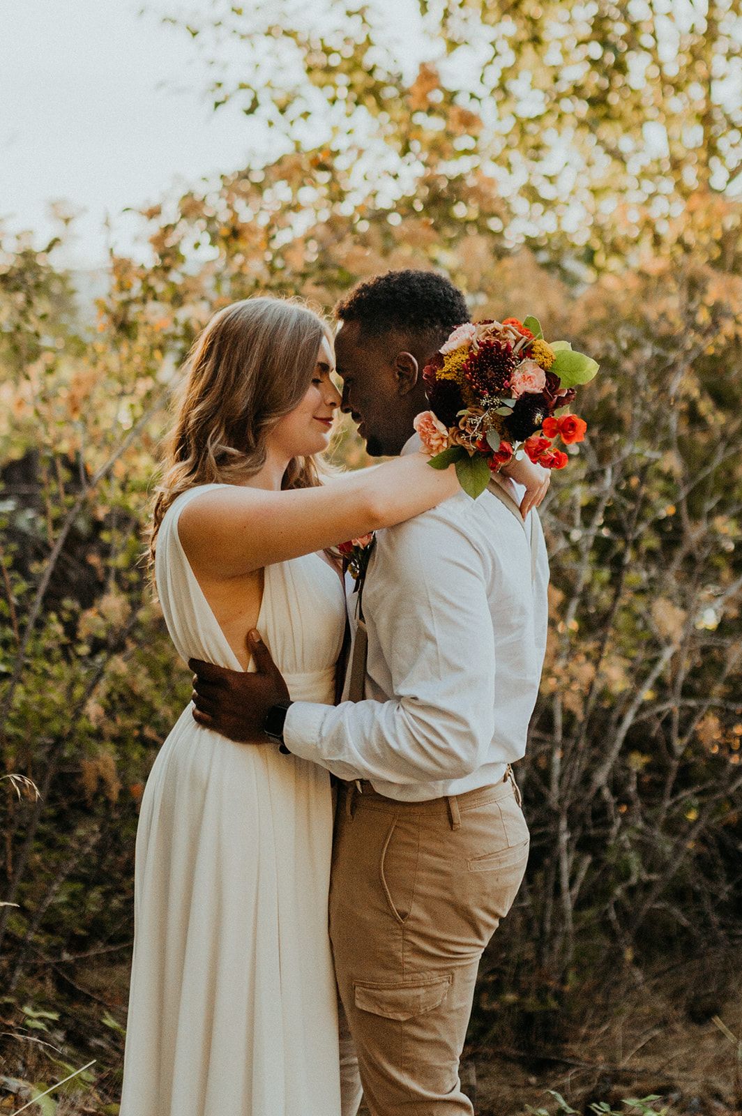 Couple in wedding attire sharing intimate moment among autumn foliage with floral bouquet.