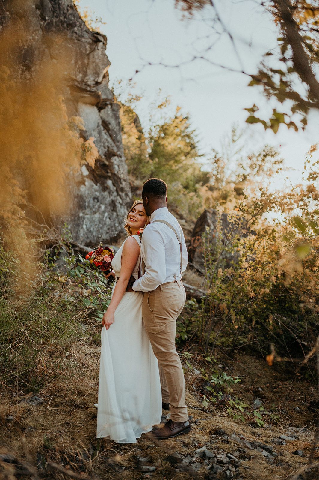 Couple embraces on a rocky hillside during golden hour sunset in a romantic outdoor photoshoot.