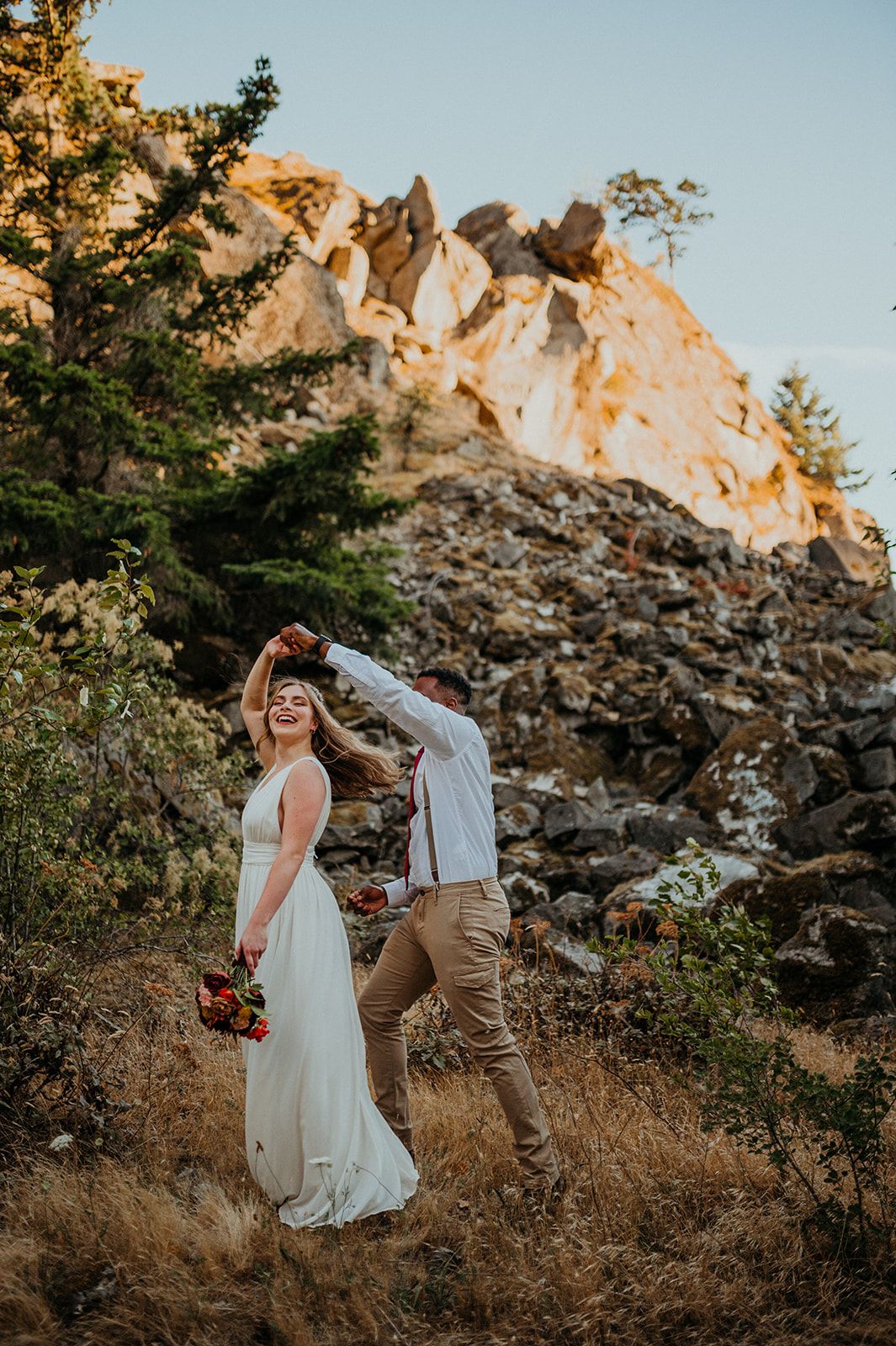 Wedding couple dances and laughs together on a mountainside trail during sunset.