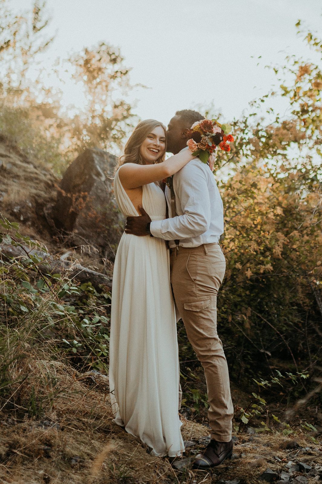 Wedding couple embracing on rocky cliff with autumn foliage in background during golden hour.