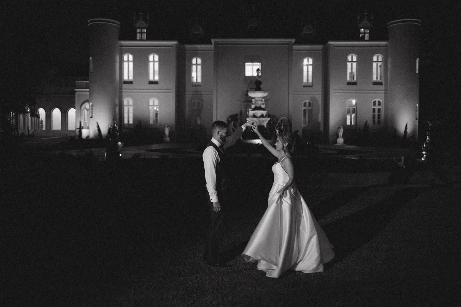 Romantic black and white photo of couple dancing outside illuminated mansion at night.