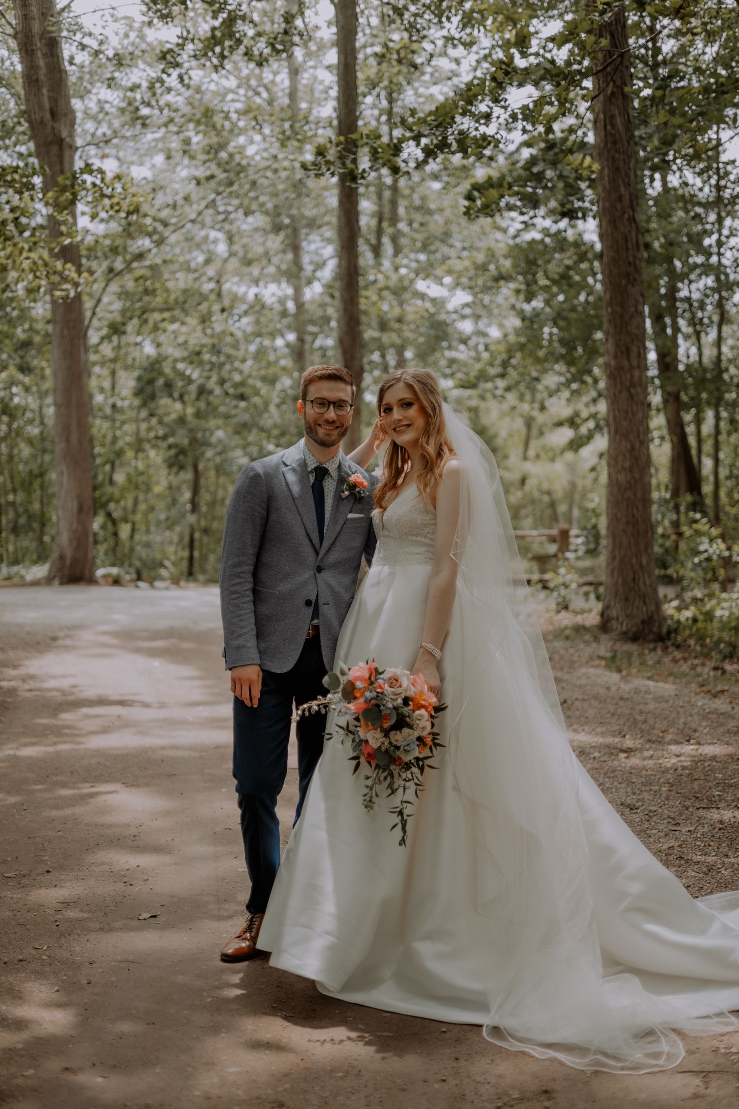 a bride and groom pose for a photo on a shaded path through the woods