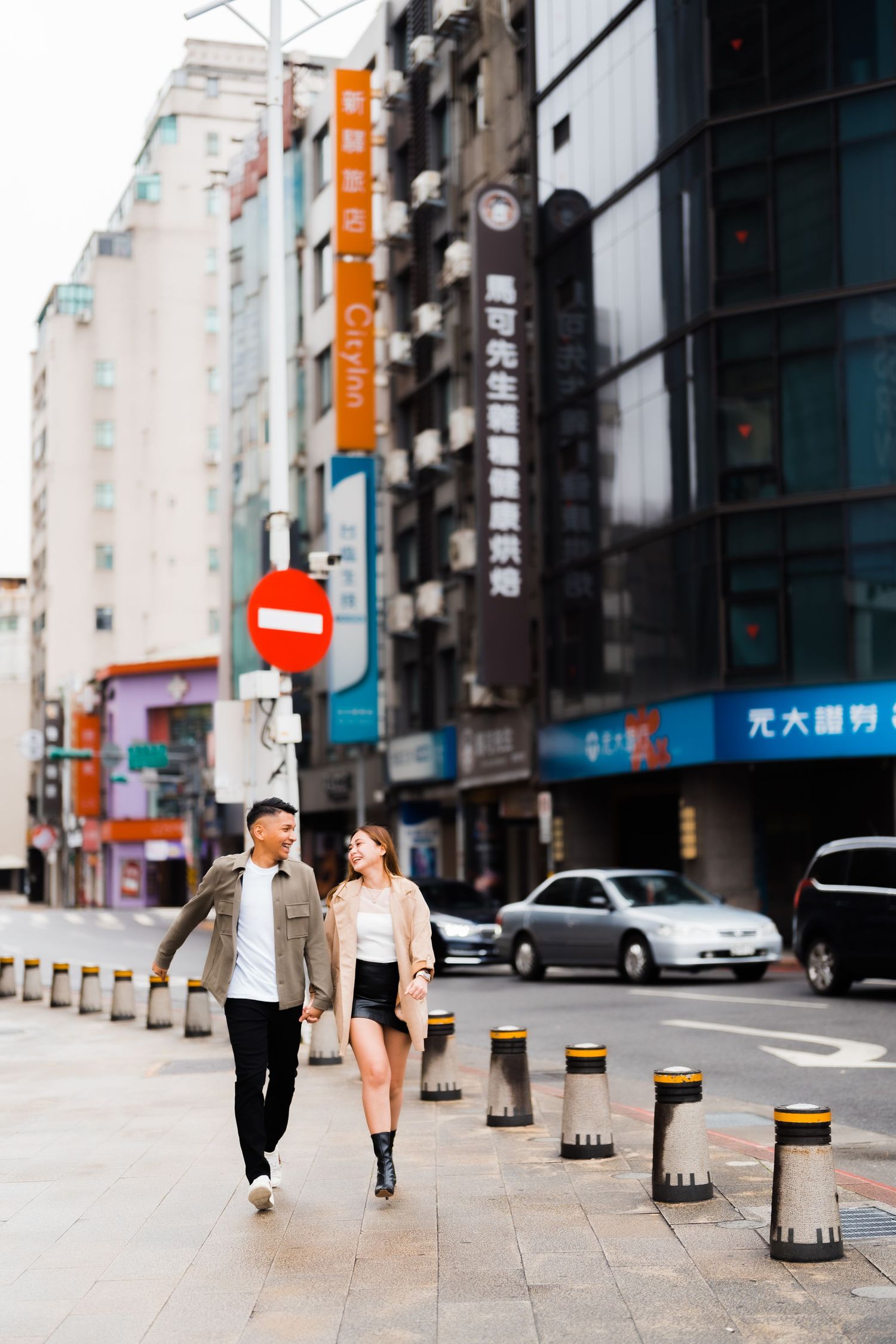 Couple walking along urban sidewalk with bollards and city buildings in background.