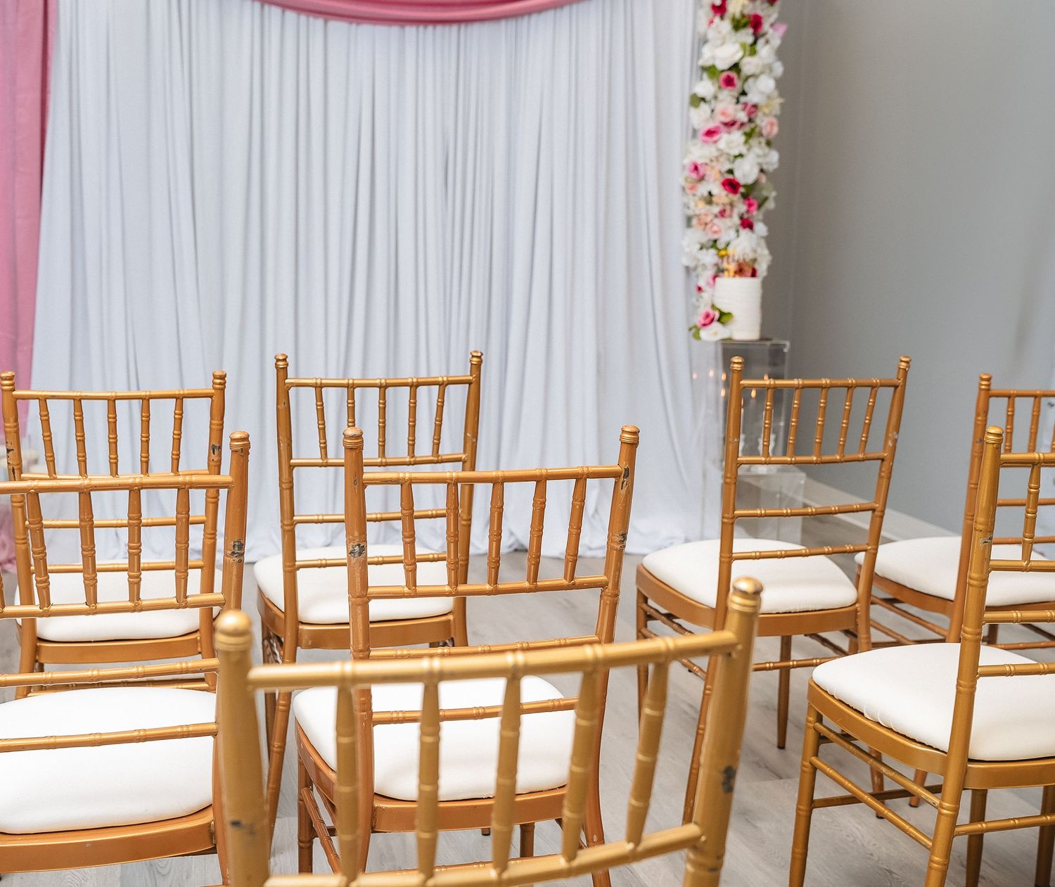 Wedding ceremony setup with wooden chiavari chairs arranged in rows against a white draped backdrop.