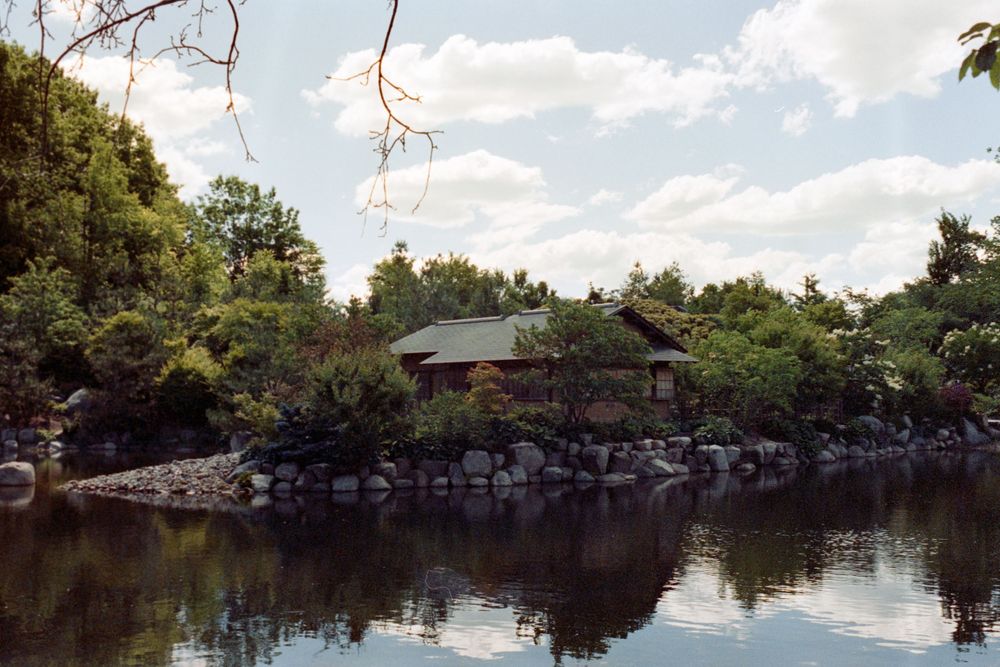 Japanese garden pavilion sits on rocky shoreline of serene pond under blue sky with puffy clouds.