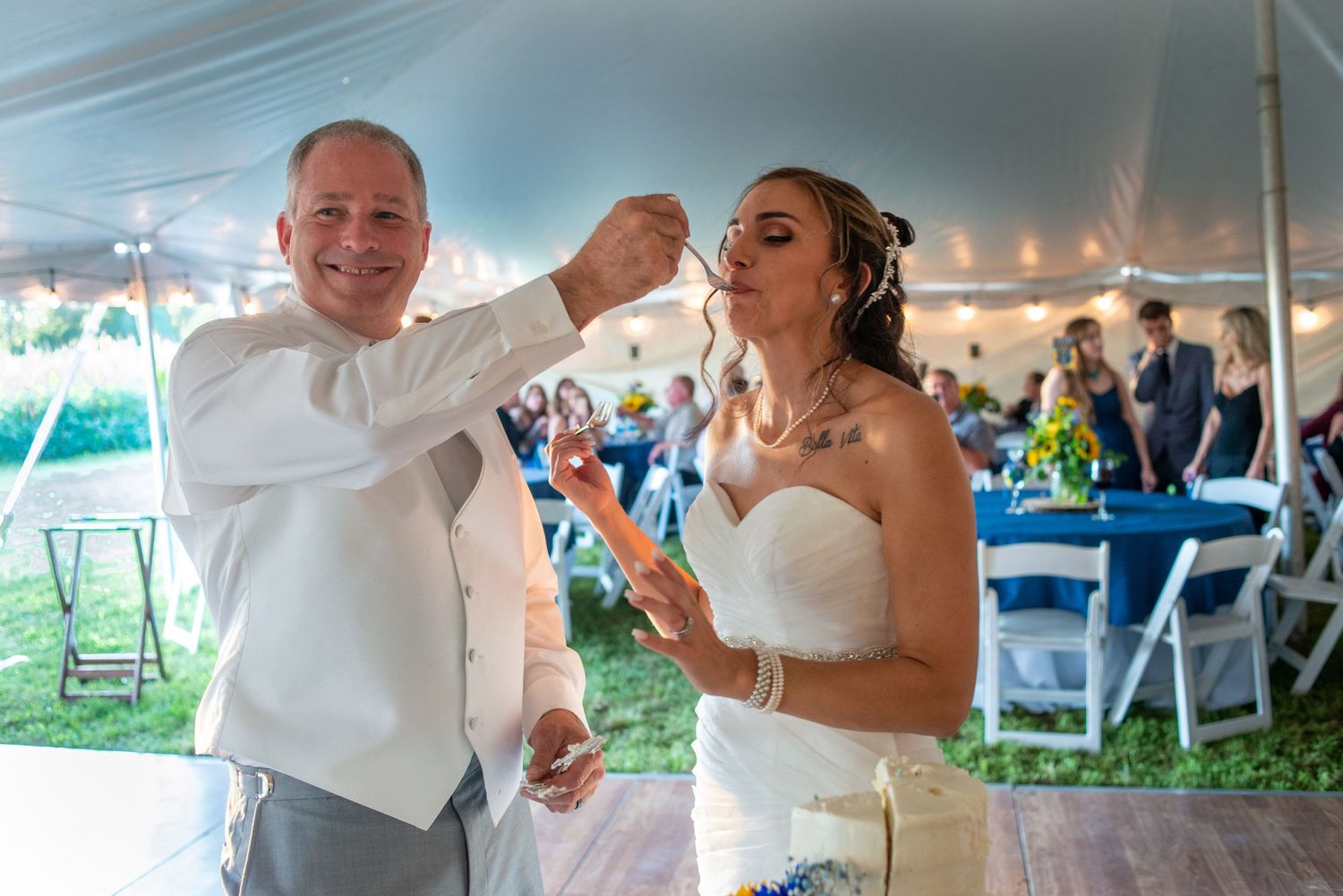 Groom playfully feeds cake to bride under a white reception tent.