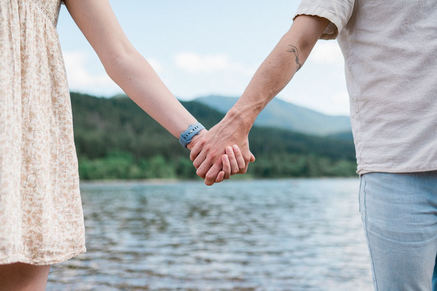 Two people hold hands by a serene lake with mountains in the background.