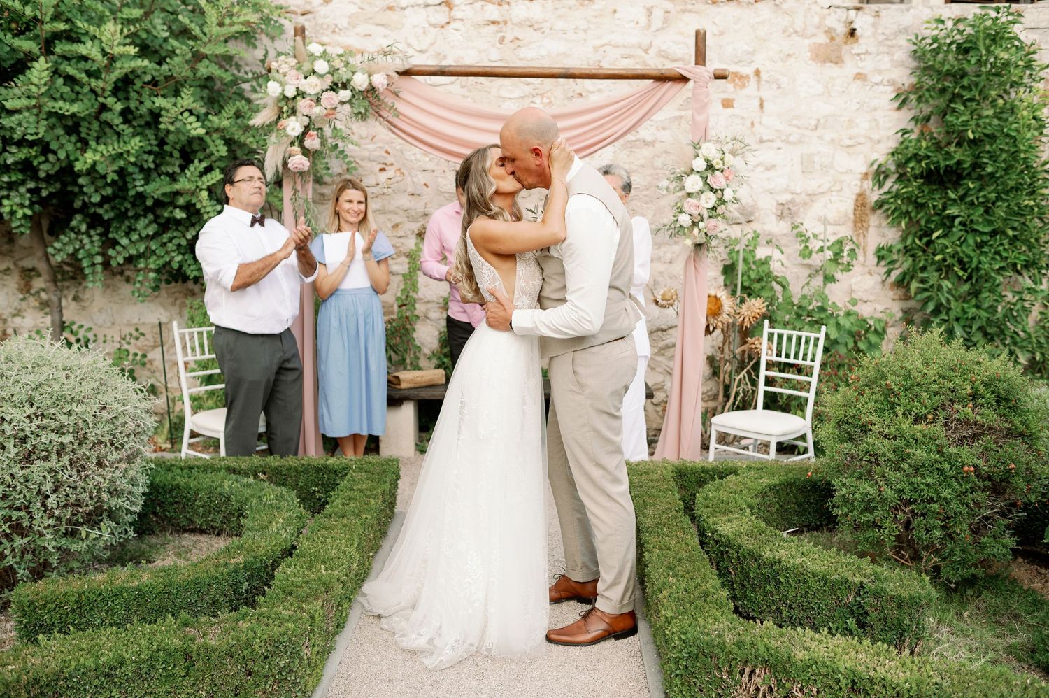A joyful wedding embrace in a garden setting with geometric hedges and stone wall backdrop.