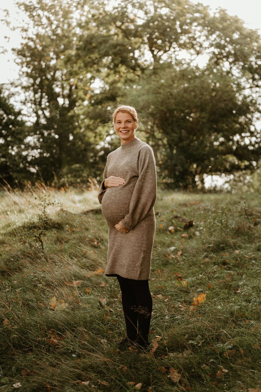 A person in a cozy sweater dress stands in a natural outdoor setting surrounded by autumn foliage.