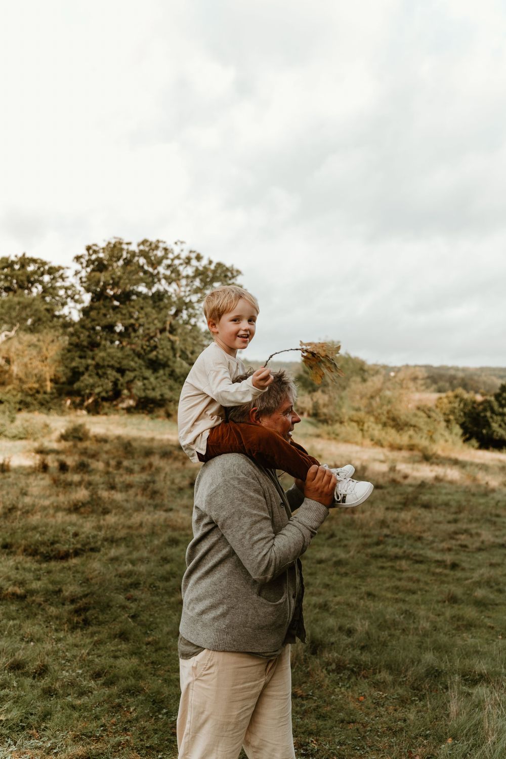 A sweet autumn moment with a child riding on shoulders amongst trees and grass on a cloudy day.
