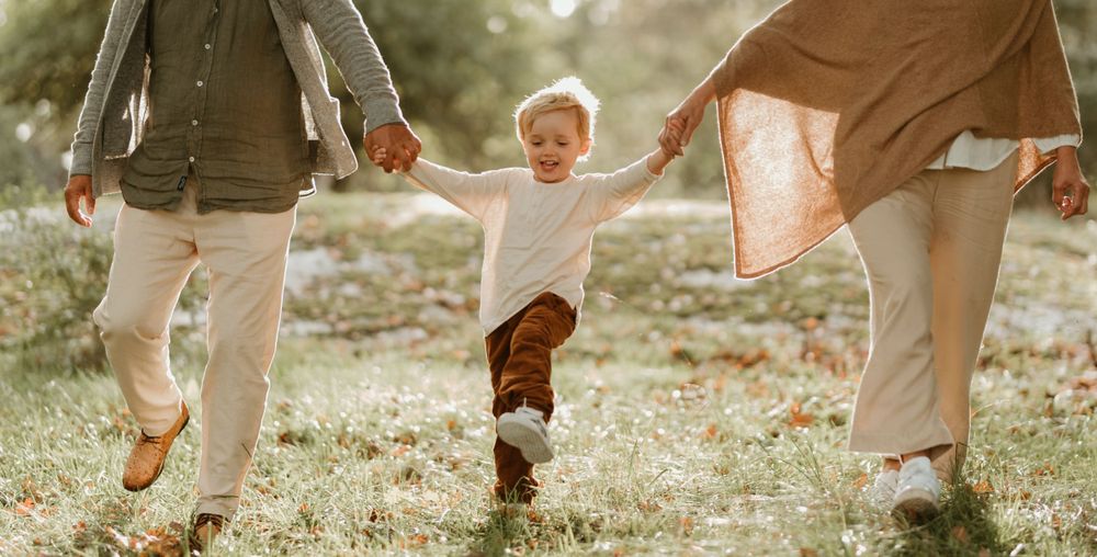 A child holds hands with two adults while jumping along a sunlit path.