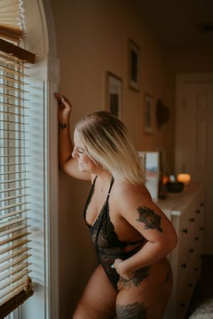 Intimate boudoir photo of a person in black lace lingerie standing by a window with visible tattoos.