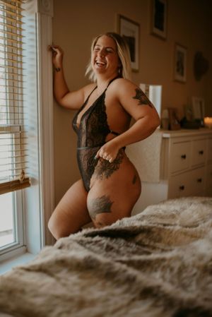 Natural light boudoir portrait capturing joyful expression and body art by window.