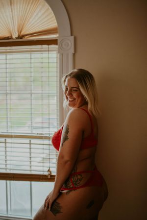 Natural light portrait by arched window wearing red lingerie with visible tattoos.