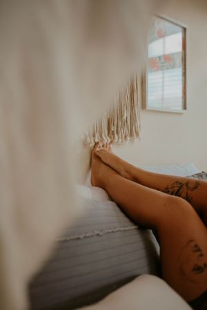 Tattooed legs resting on white bedsheets with a framed artwork visible on the wall in soft, natural lighting.