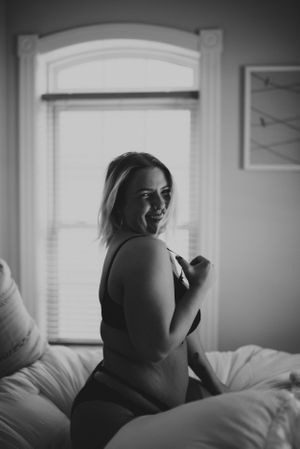 Black and white boudoir photo of a woman sitting on a bed in front of a bright window, captured in natural light.