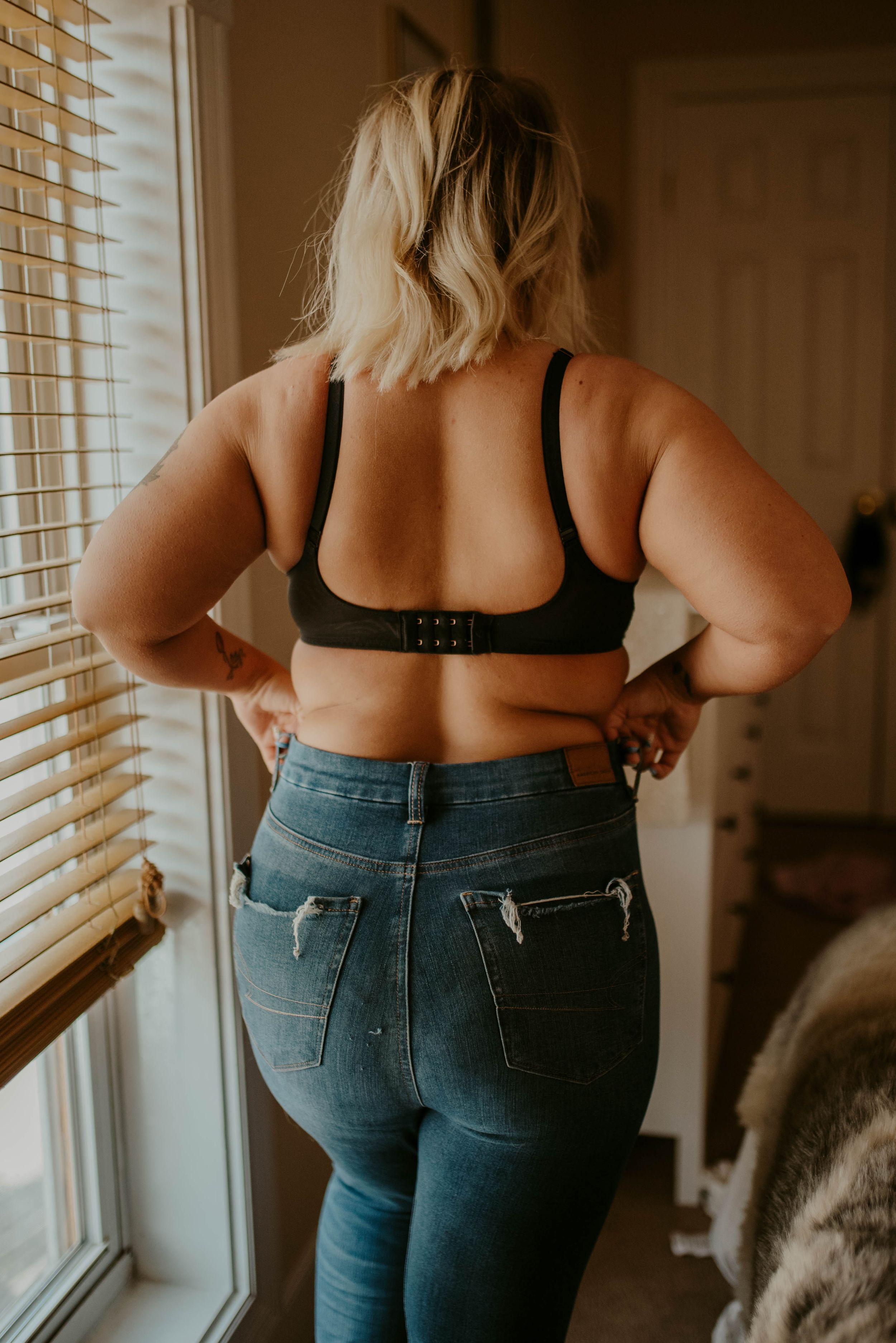 Back view of person wearing black sports bra and high-waisted blue jeans standing by window with venetian blinds.