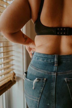 Close-up view of person wearing high-waisted distressed jeans and black bra top standing near window blinds.