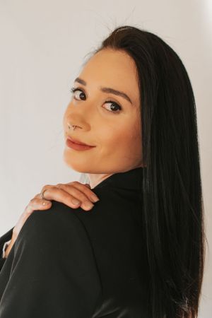 Over-the-shoulder pose in black blazer against white background with natural makeup.