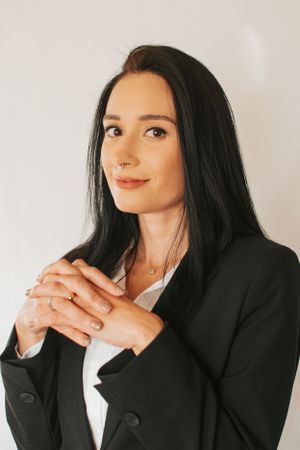 Professional headshot with clasped hands wearing black blazer and delicate necklace.
