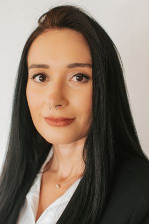 Close-up professional portrait in black blazer with minimalist jewelry against white background.
