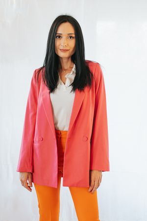 Professional photoshoot featuring bright coral blazer with orange pants and white top against light background.