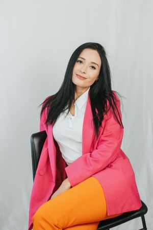 Stylish seated pose in coral blazer and orange pants on black chair against white background.