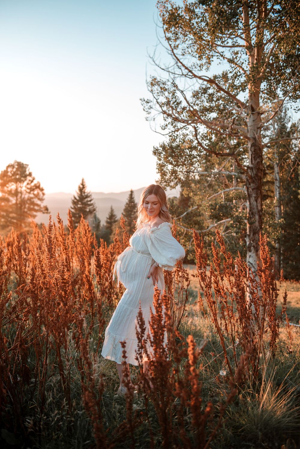 A white dress flows in a golden sunset field surrounded by tall red wildflowers and pine trees.