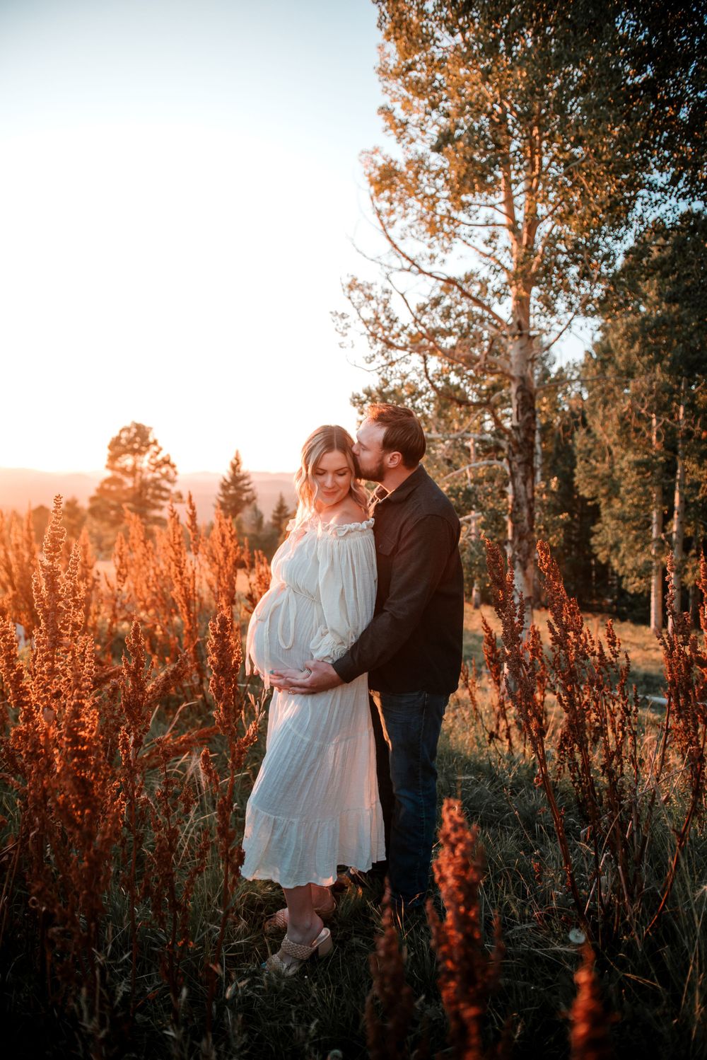 Couple poses together in rustic outdoor setting with red flowers and pine trees at sunset.