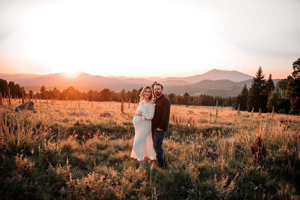 A couple embraces in a golden mountain meadow at sunset with evergreen trees in the background.