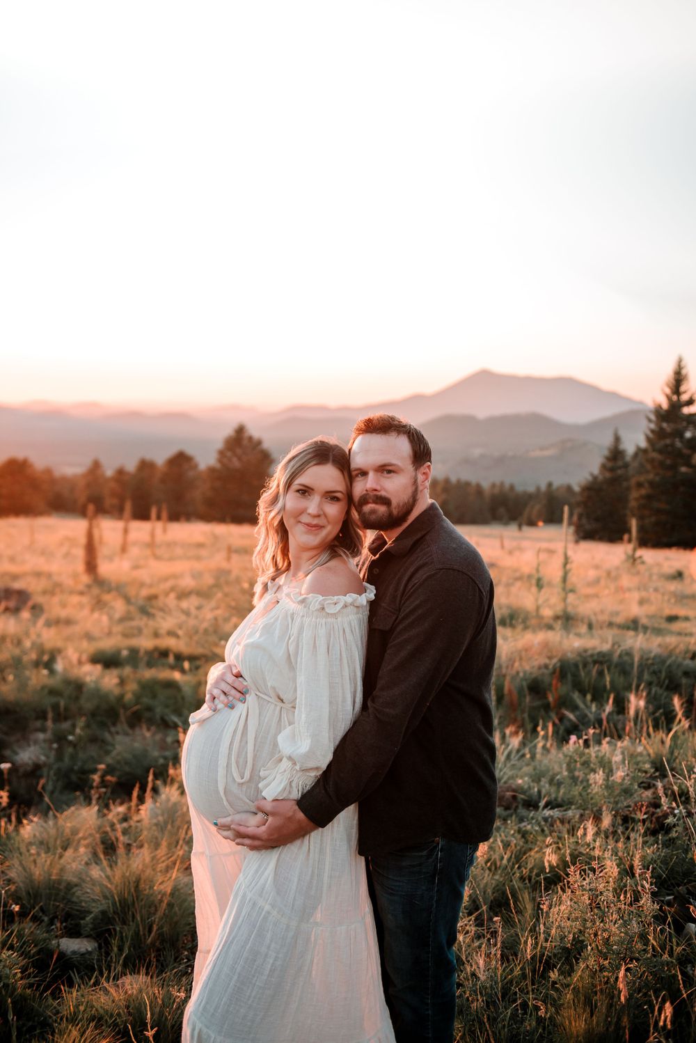 A maternity photo of a couple in a mountain field during golden hour sunset.