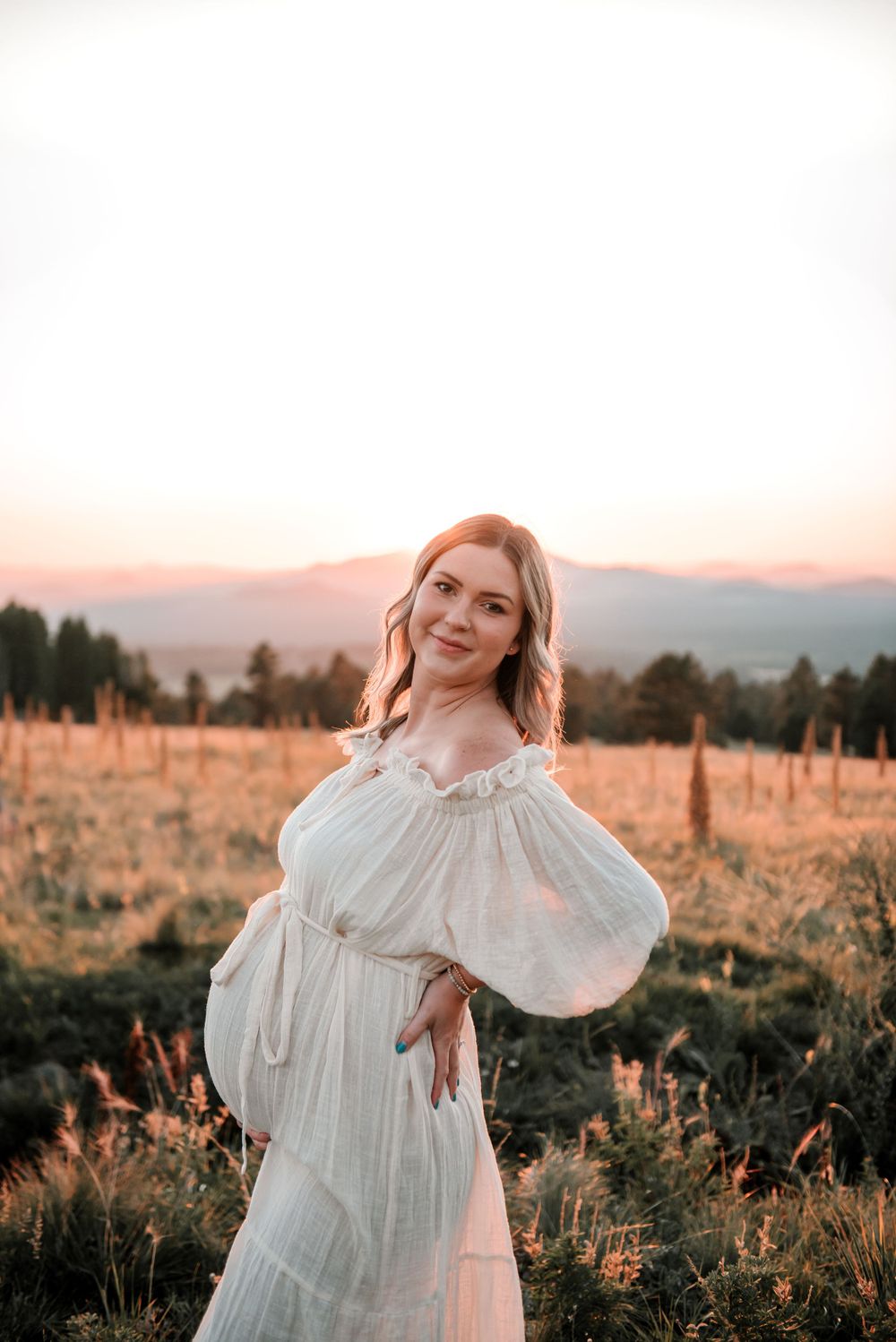 Woman in white flowing dress poses in mountain meadow during sunset.