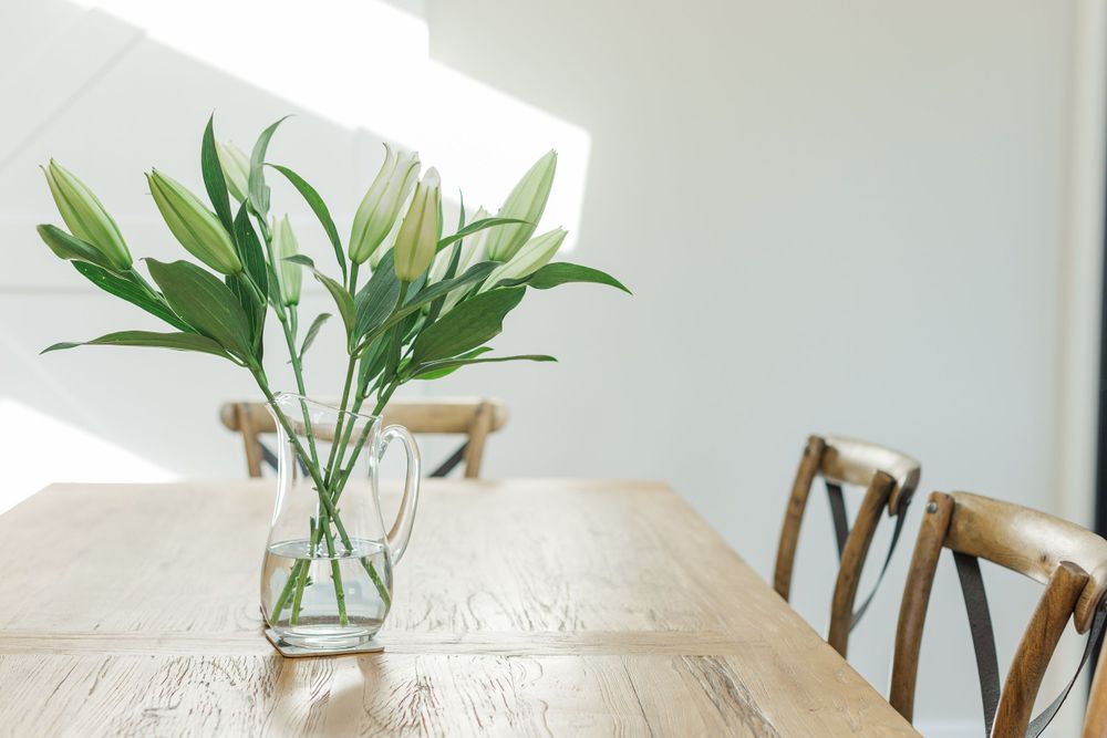 Fresh white lilies in a glass vase on a wooden dining table with chairs.