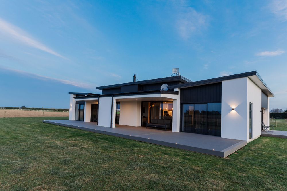 Modern white house exterior with large windows and covered patio at dusk.
