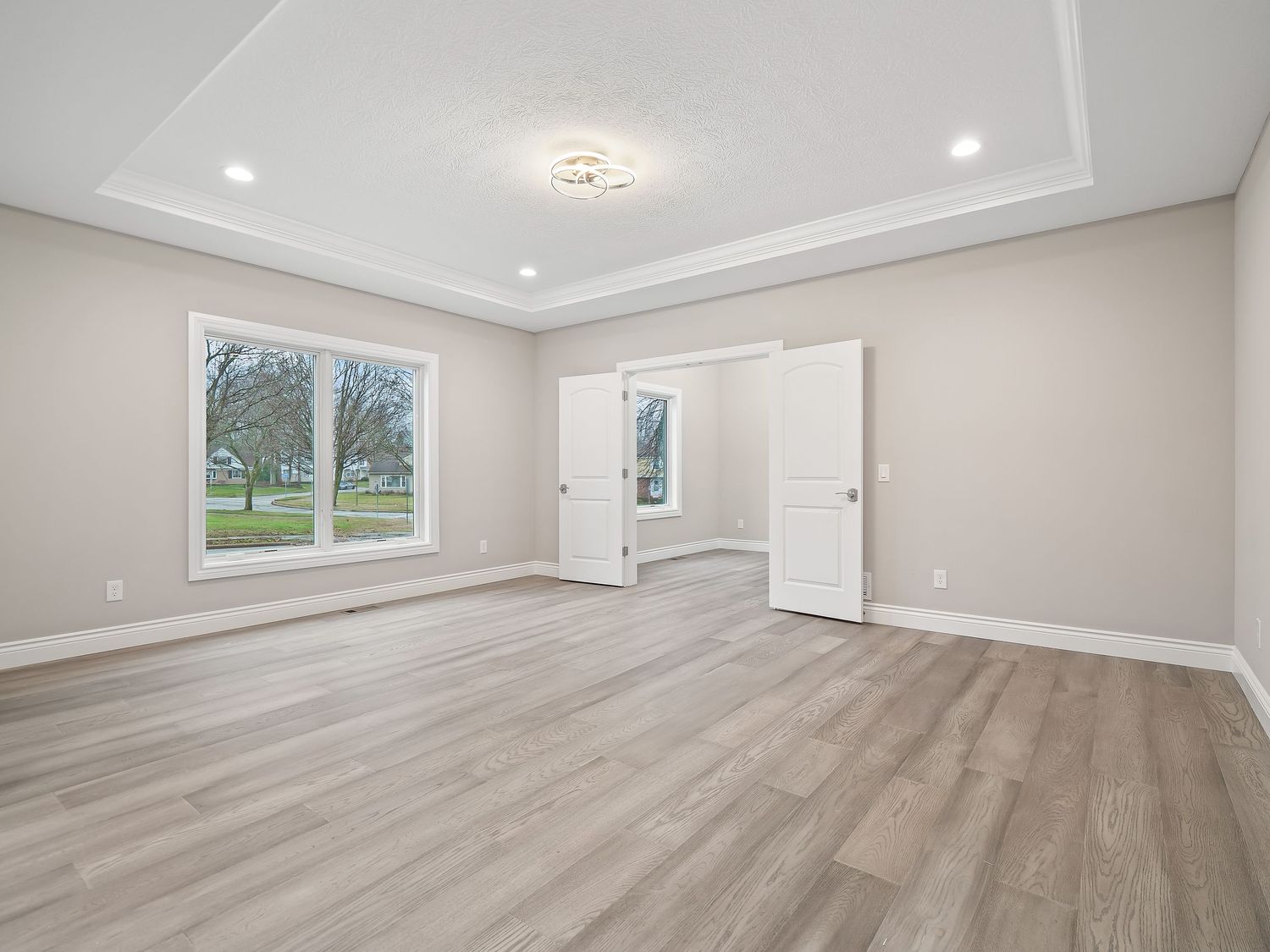 Empty room with gray wood flooring, tray ceiling, and large windows overlooking nature.