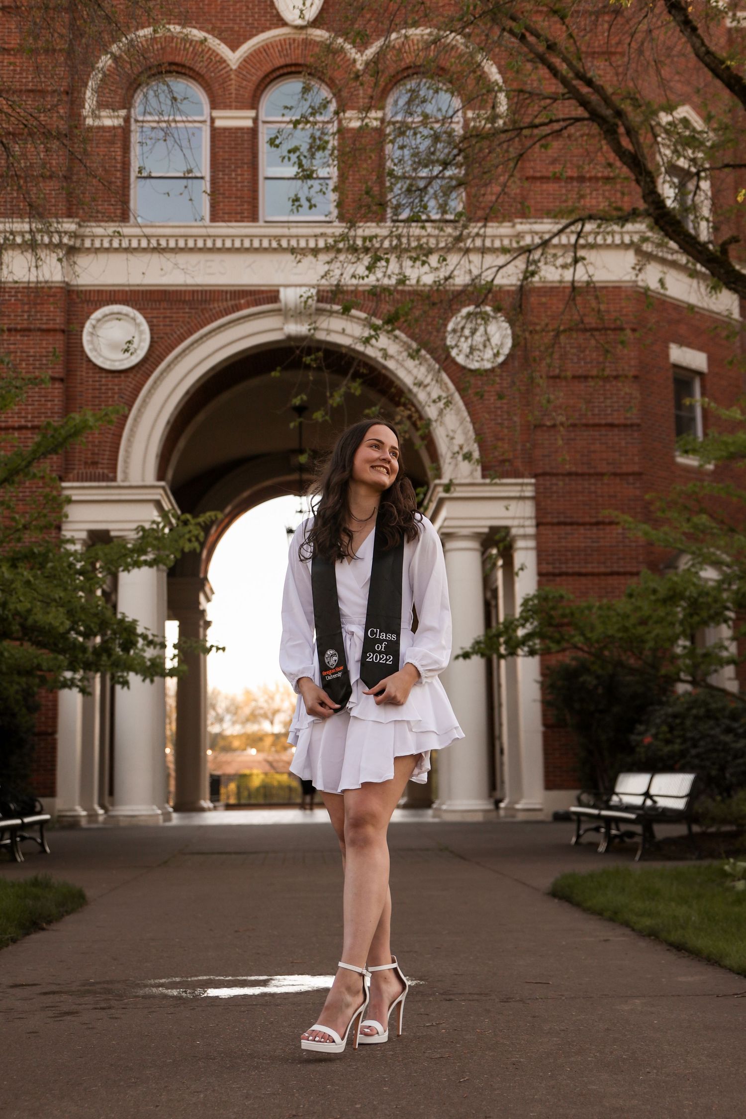 Person in white dress and blazer poses in front of historic brick building with arched entrance.