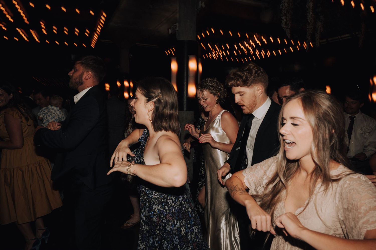 Wedding reception guests dancing and laughing together under moody lighting and string lights.