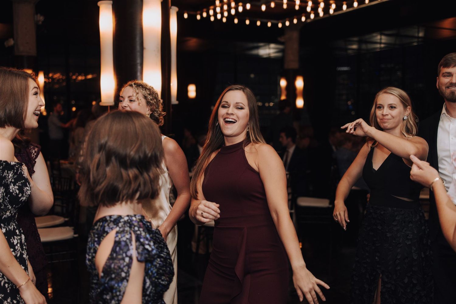 Group of party guests dancing and laughing together under festive string lights at an event.