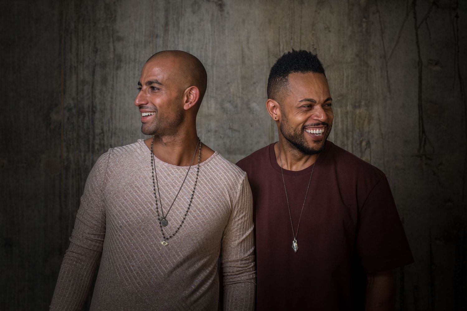 Two men in casual sweaters pose together against textured wall in portrait lighting.
