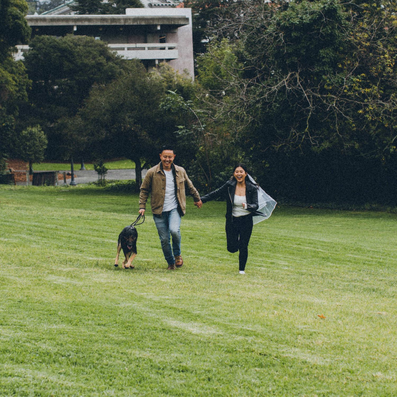 A couple runs playfully across a grass field with their black dog on a cloudy day.