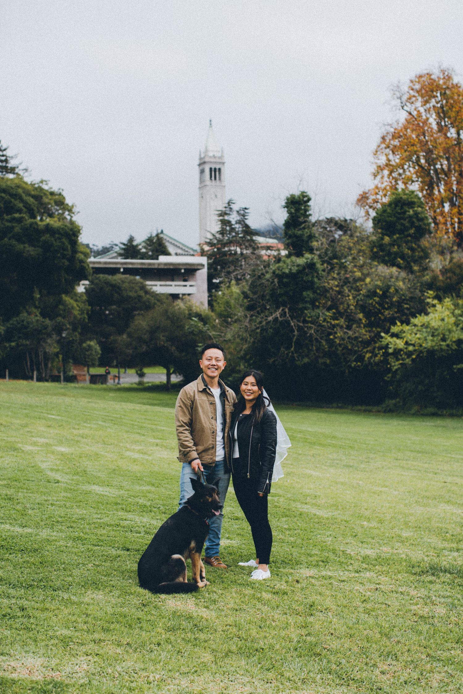 A couple and their black dog pose together on a grassy hill with a bell tower in the background.