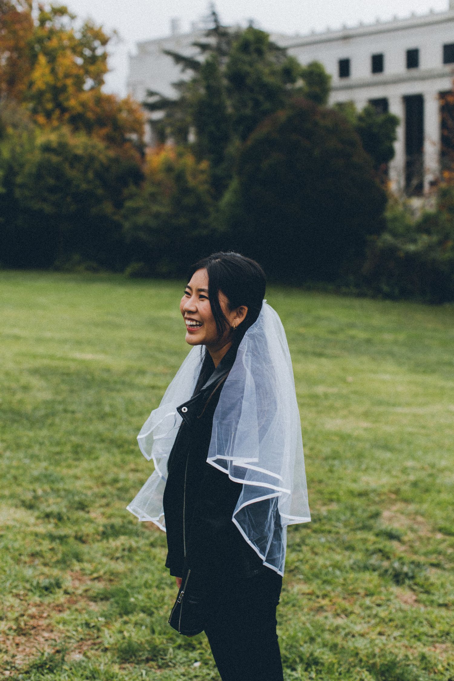 A person in a black outfit and white veil smiles joyfully on a grassy lawn.