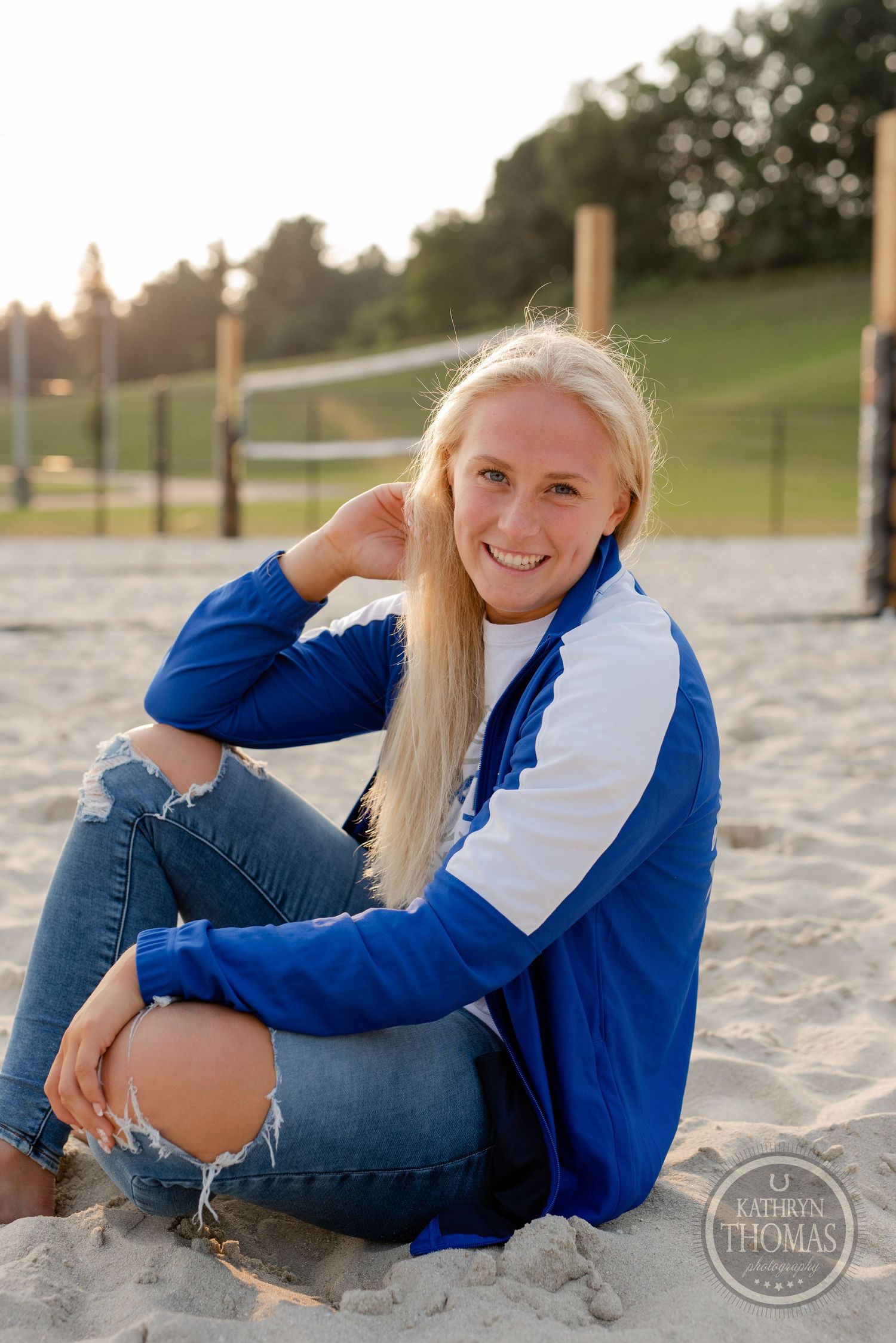 Athlete in blue and white jacket sits on sandy volleyball court at sunset.