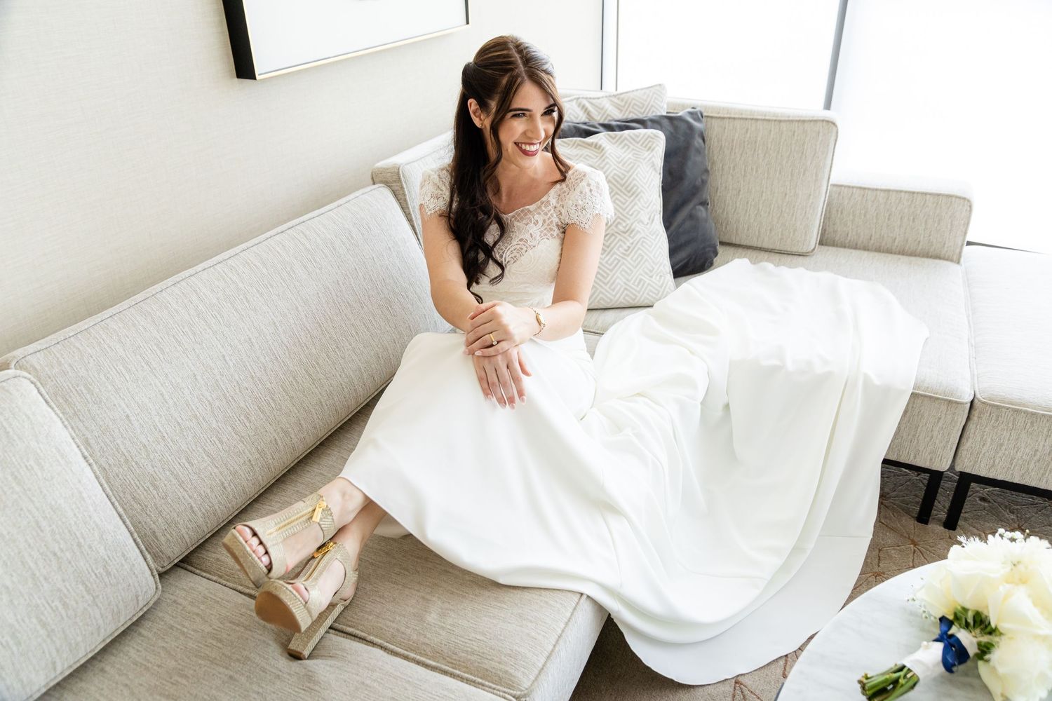Bride in classic white dress relaxing on light-colored couch with bouquet nearby.