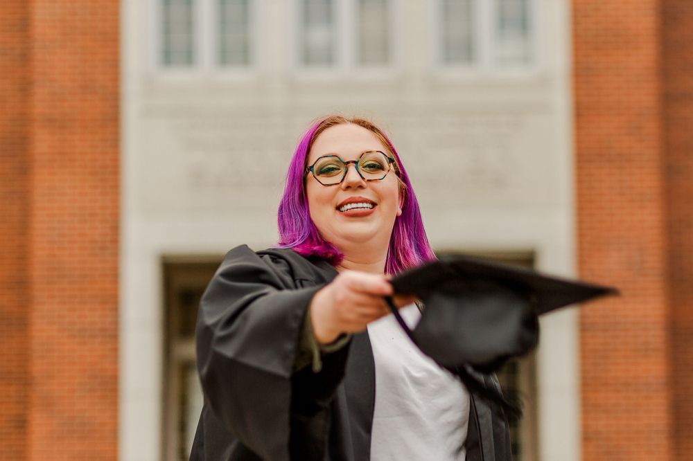 Graduate adjusts graduation cap while posing in front of academic building.