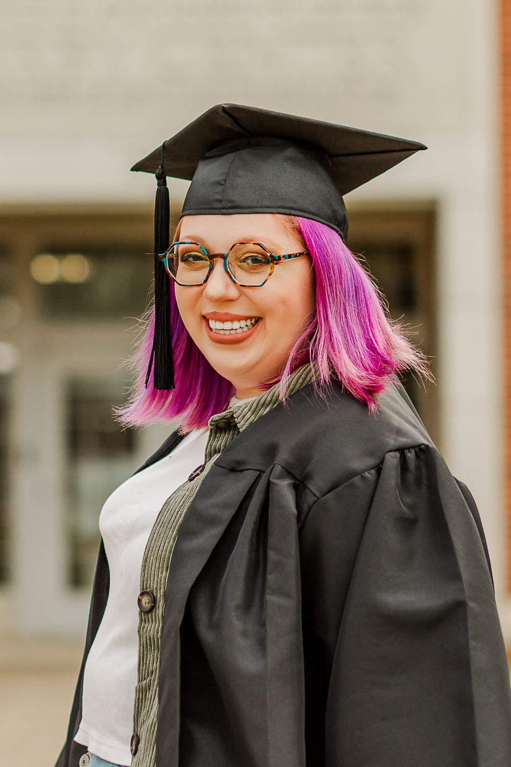 Graduate in black cap and gown with vibrant pink hair and glasses poses for portrait on campus.